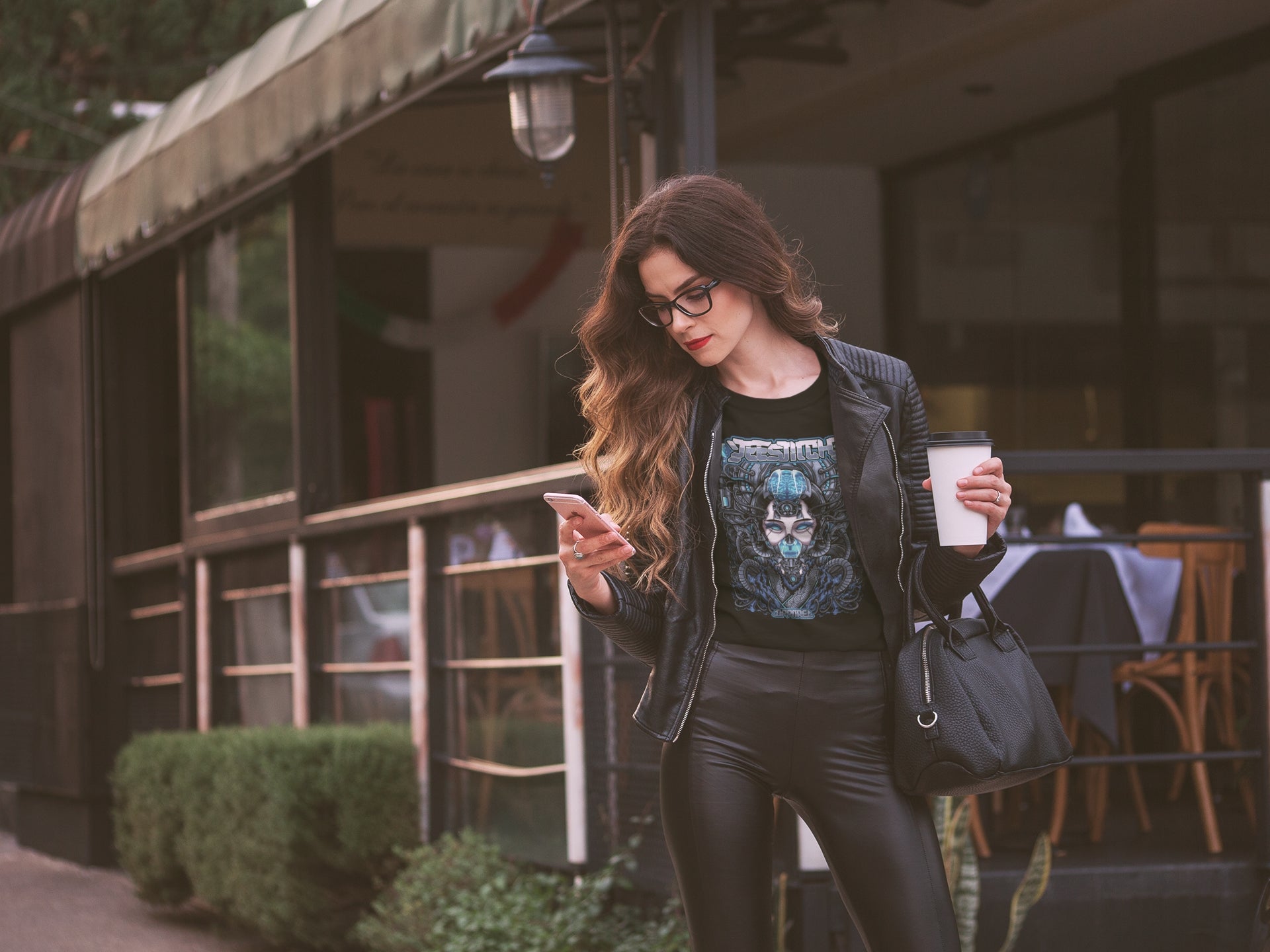 Woman standing outside holding a phone and a coffee cup, wearing a black leather jacket and graphic t-shirt, with long wavy hair and glasses.