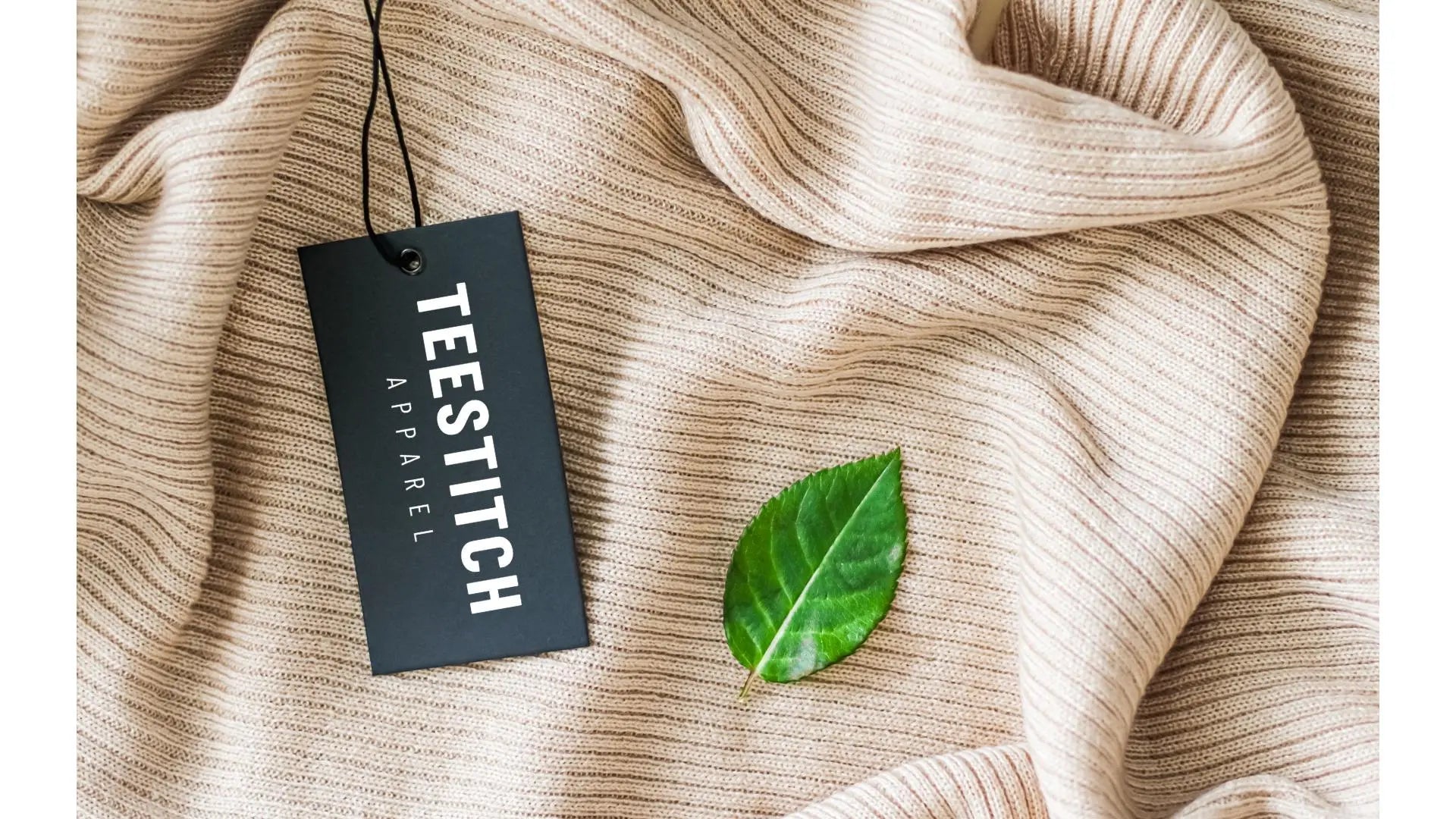 Black TeeStitch Apparel clothing tag resting on a soft beige knit fabric with a green leaf, symbolizing sustainable and ethical fashion.