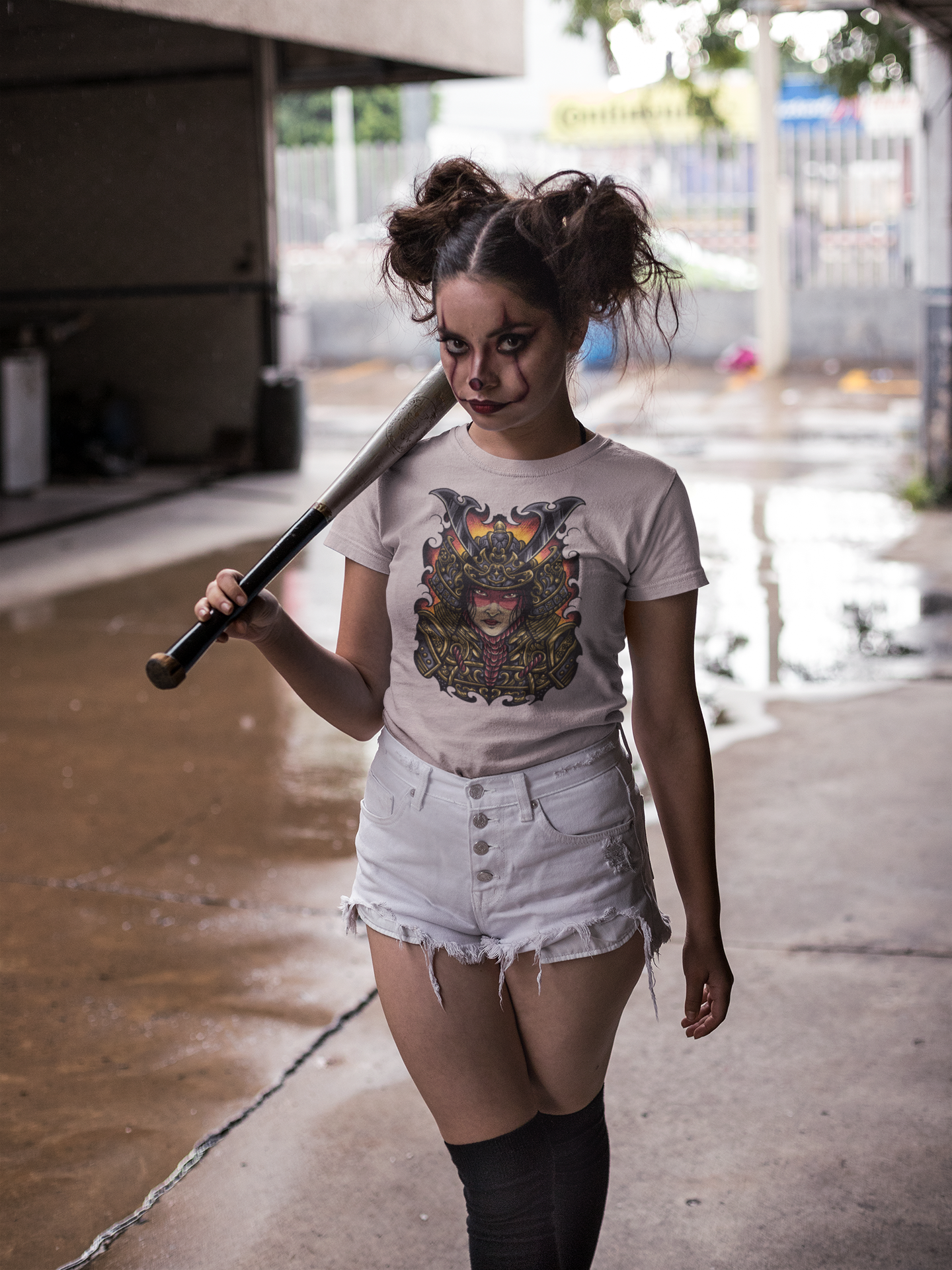 A woman stands in a wet outdoor alley holding a baseball bat over her shoulder. She is wearing a light pink graphic T-shirt with a Lady Ronin exclusive design, white denim shorts, and black knee-high socks. TeeStitch Apparel