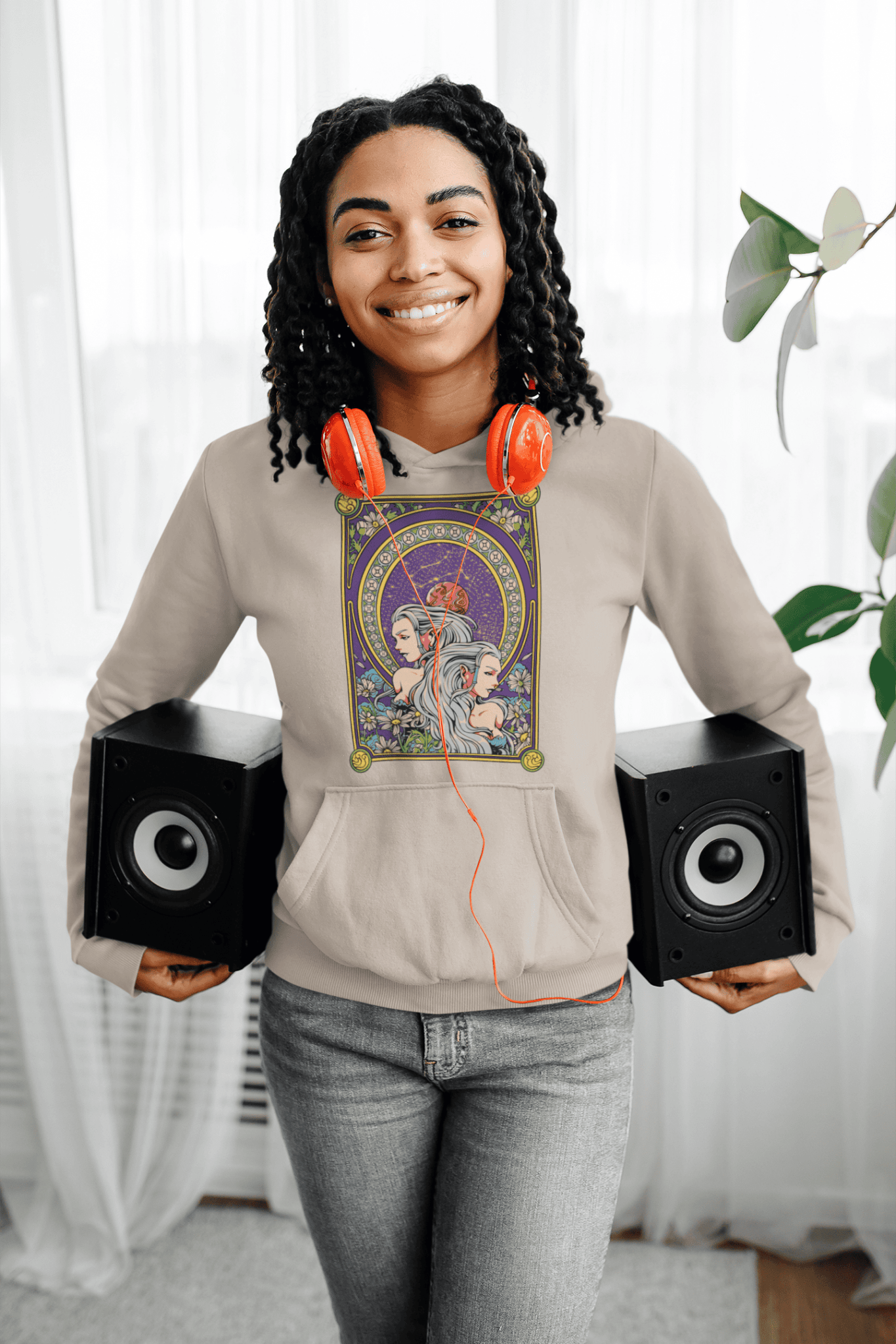 Person wearing a beige sweatshirt with a graphic design, holding speakers, in a room with curtains and a plant.