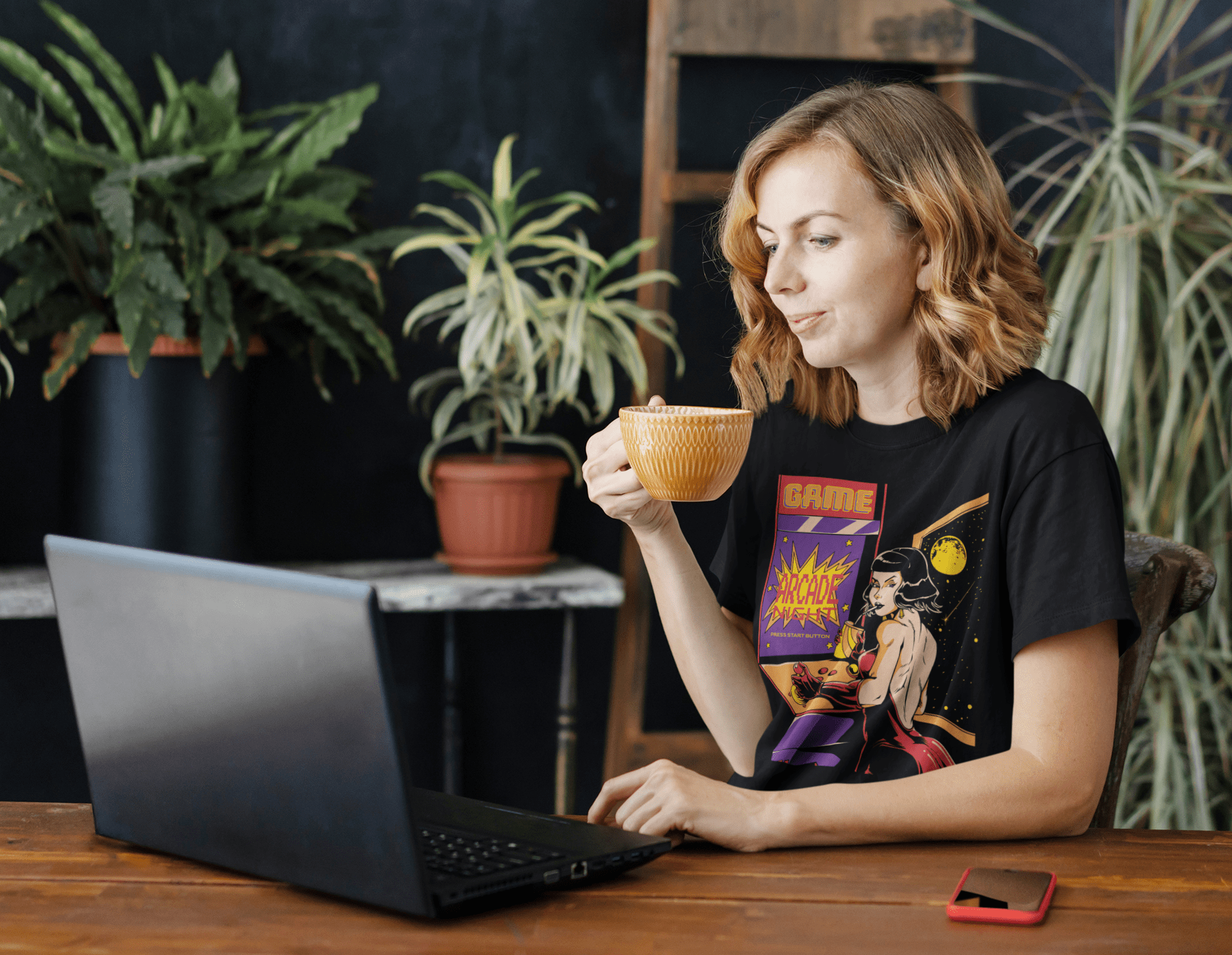 women sitting at her laptop working while drinking coffee wearing a TeeStitch Apparel Arcade Night custom designed t-shirt